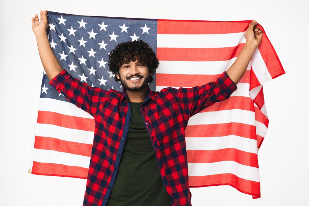 A young Hindi-American male wearing casual union-made apparel and holding an American flag.